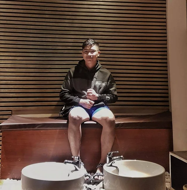 Rechcel in a black hoodie seated on a bench against a wooden slat wall